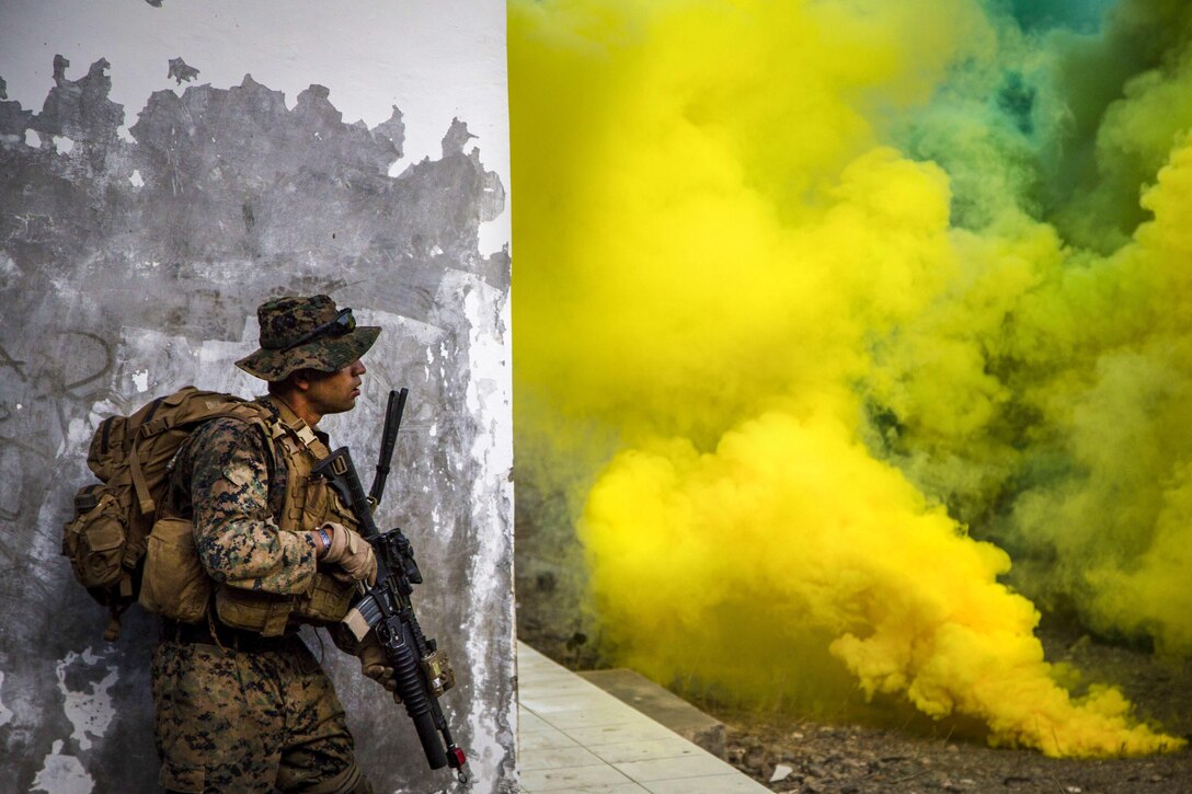 A Marine stands against a wall with a weapon, looking out at yellow and green smoke from smoke bombs.