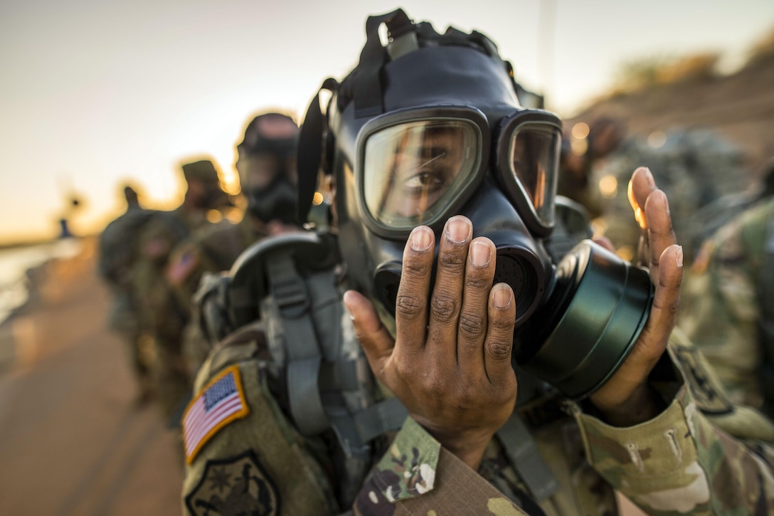 A soldier holds her hands over the canisters of a gas mask she is wearing.