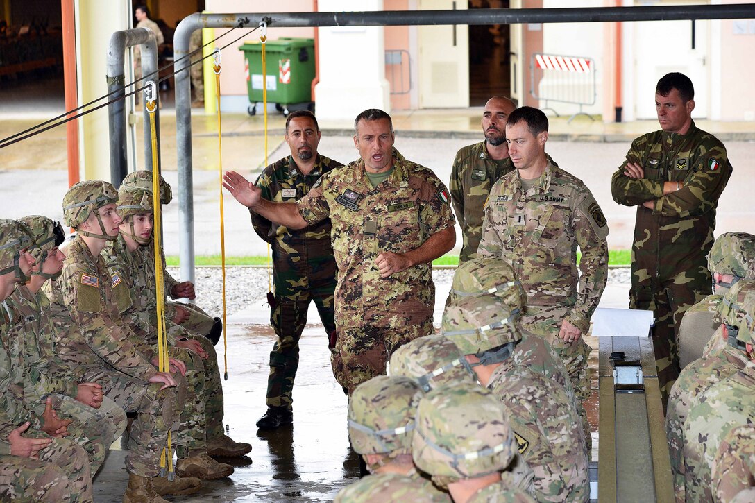A group of sittin gsoldiers listens to an Italian service member stand and talk.