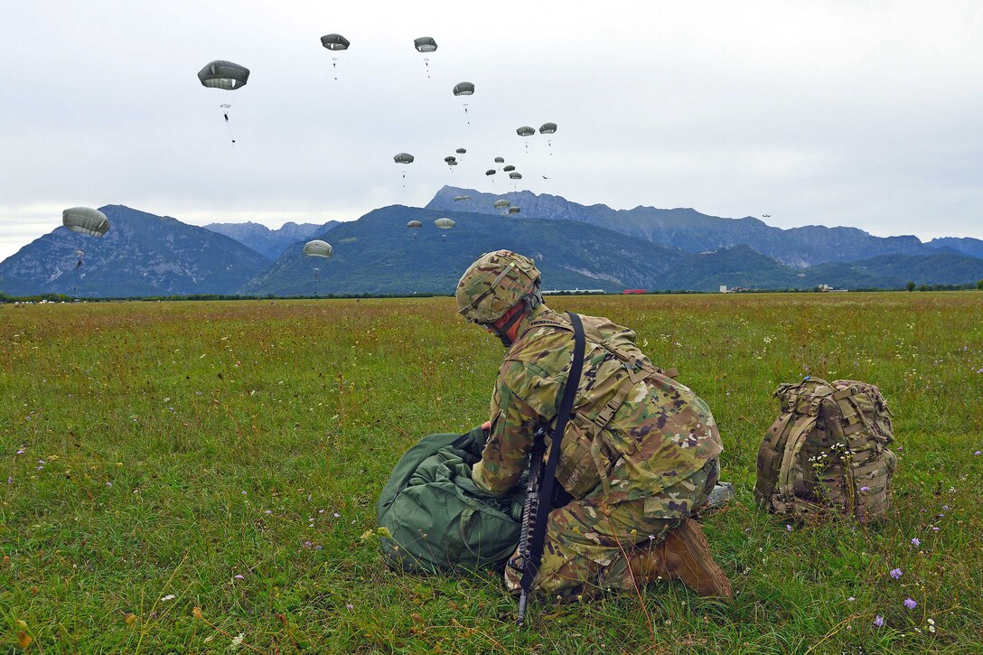 A soldier puts gear in a bag with other paratroopers drifting to the ground.
