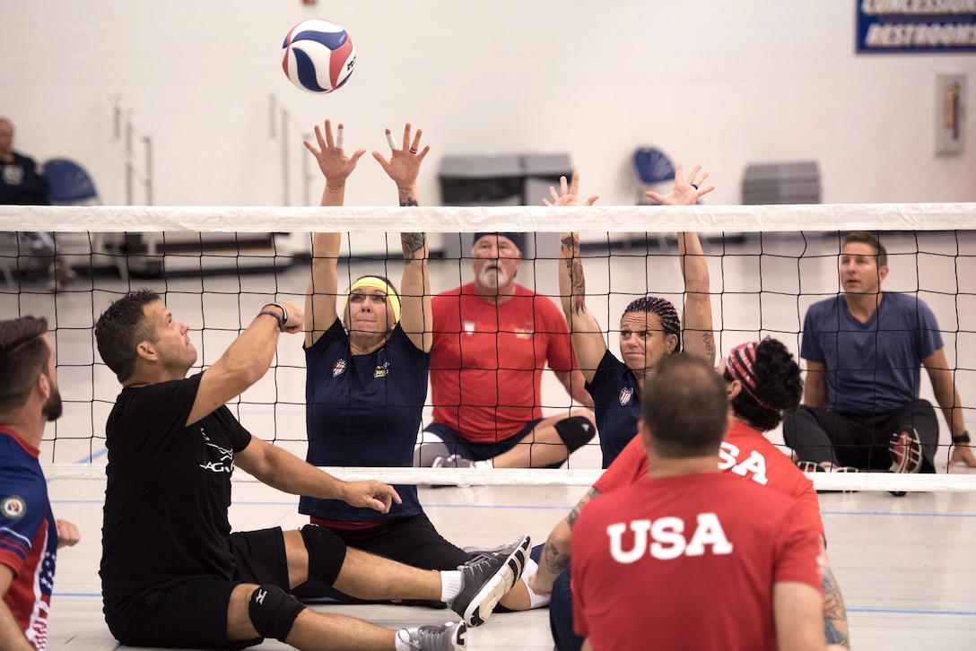 A player sits on the ground and reaches up towards a volleyball going over a net.