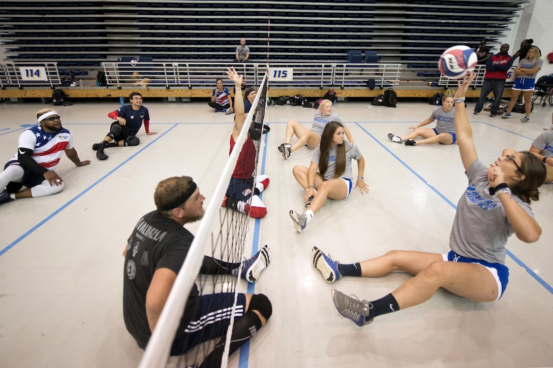 Two teams of players sit on a volleyball court while one player reaches for the ball.