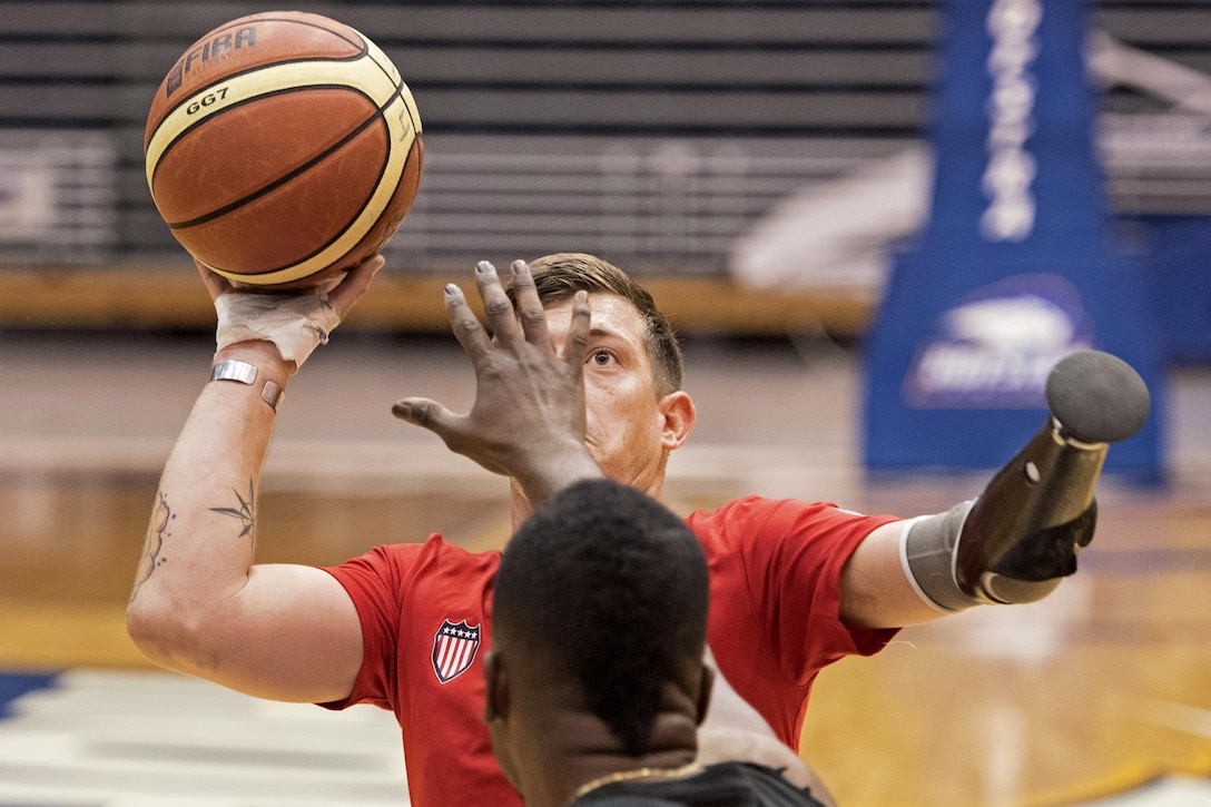 A player uses one arm to throw a basketball while another player attempts to block.