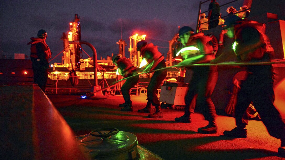 Sailors, illuminated by green lighting, pull a line on a ship against a night sky.