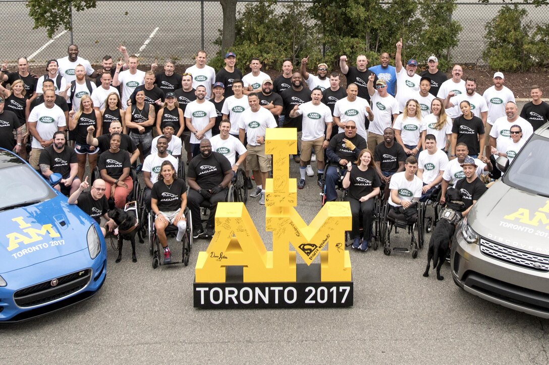 A group of Wounded Warrior athletes pose for a photo next to cars and prop saying, "I am Toronto 2017."
