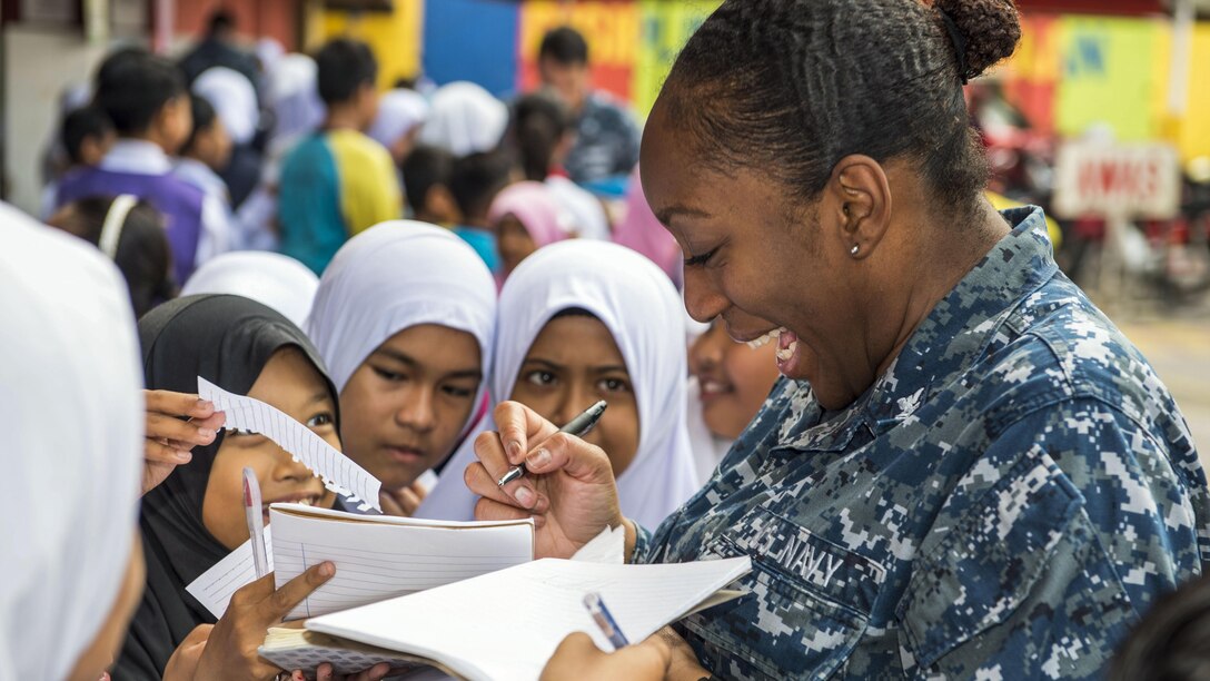 Several girls crowd around a sailor, who writes on paper they are holding out to her.
