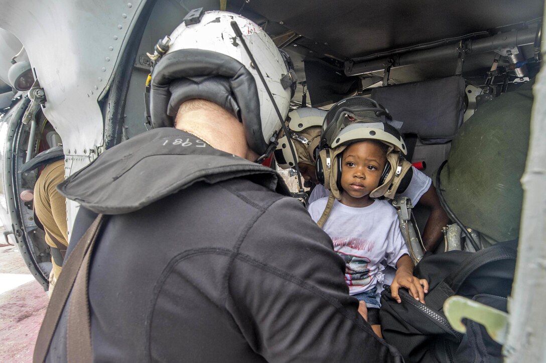 A child wearing a helmet inside a helicopter looks at a sailor.
