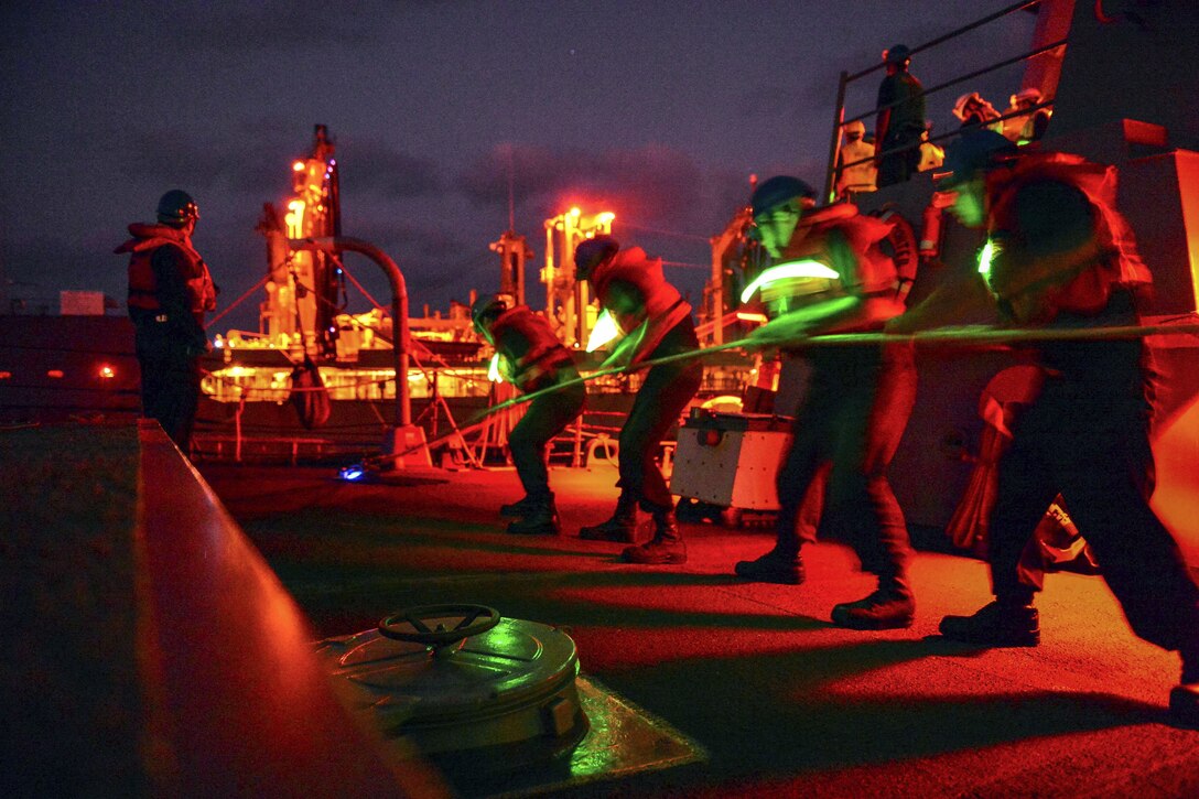 Sailors, illuminated by green lighting, pull a line on a ship against a night sky.