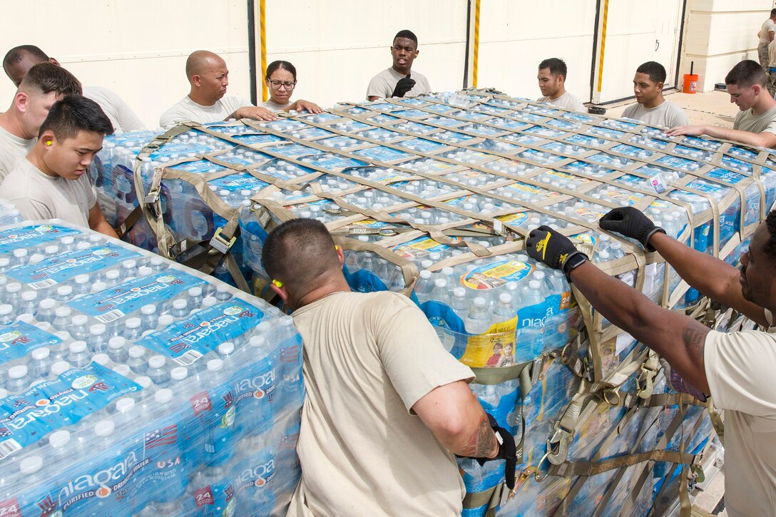 Airmen wrap and tighten cargo straps around a large pallet of water and supplies for transport.