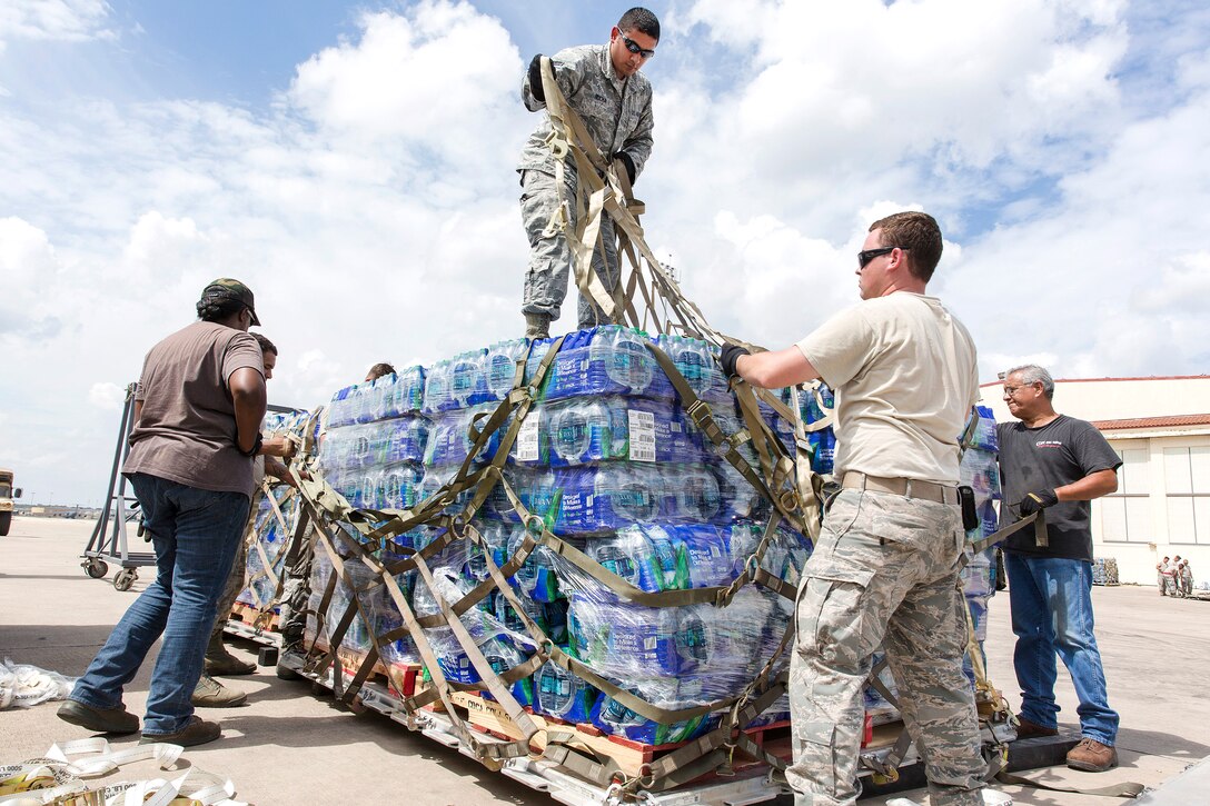 Airmen and civilians wrap and tighten cargo straps around a large pallet of water for transport.