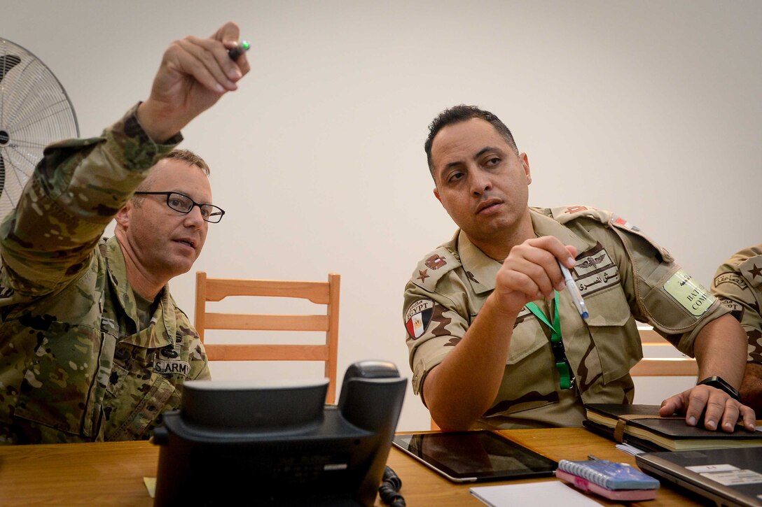 An Army colonel speaks to an Egyptian soldier while sitting at a table.