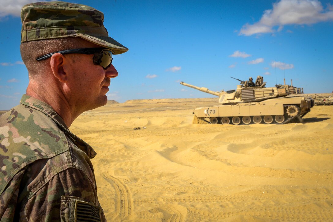 A soldier watches a tank in the sand.