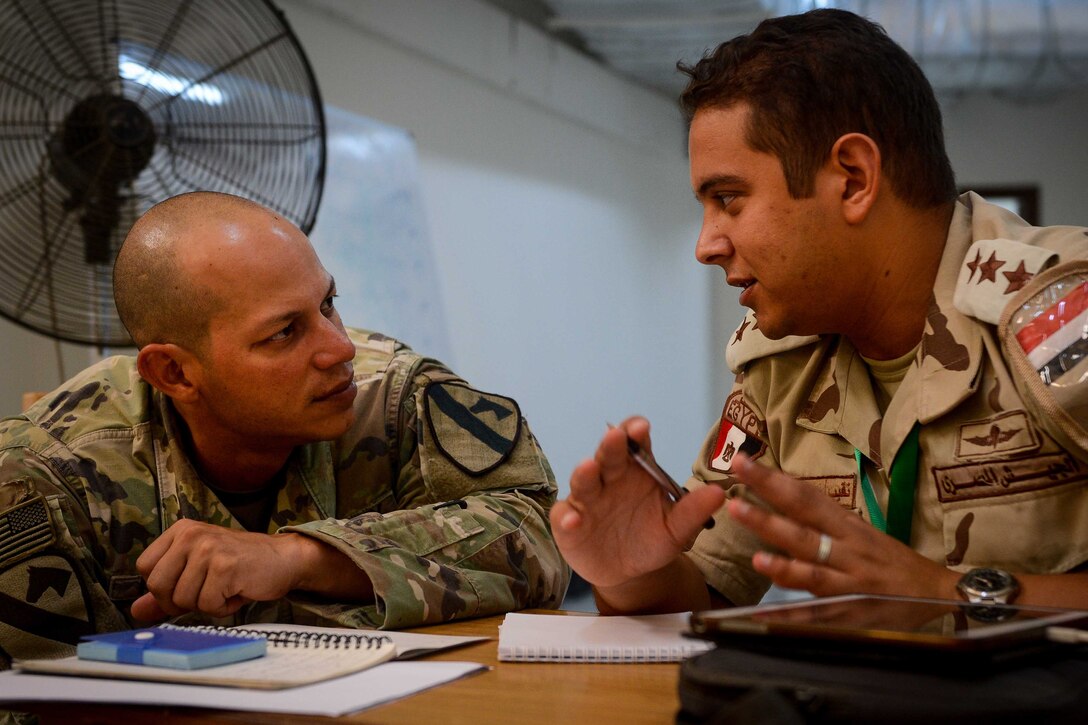 A soldier sits at a table and talks with an Egyptian soldier sitting at the table.