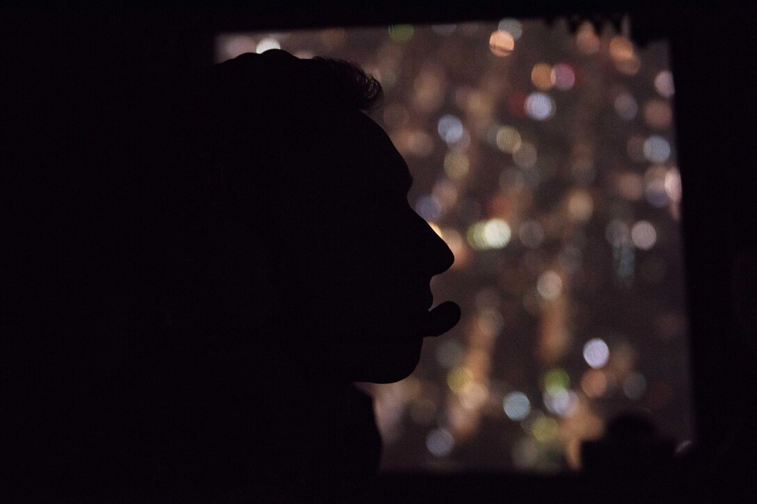 An airman is silhouetted against city lights from the aircraft window.