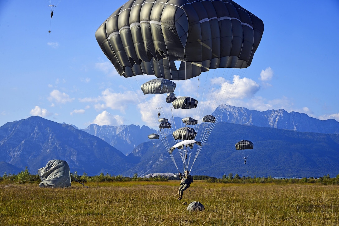 Parachutes descend to the ground, against a backdrop of mountains.