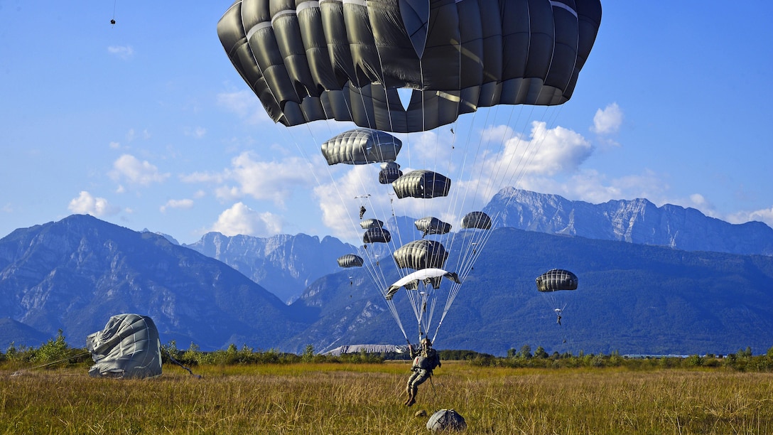 Parachutes descend to the ground, against a backdrop of mountains.