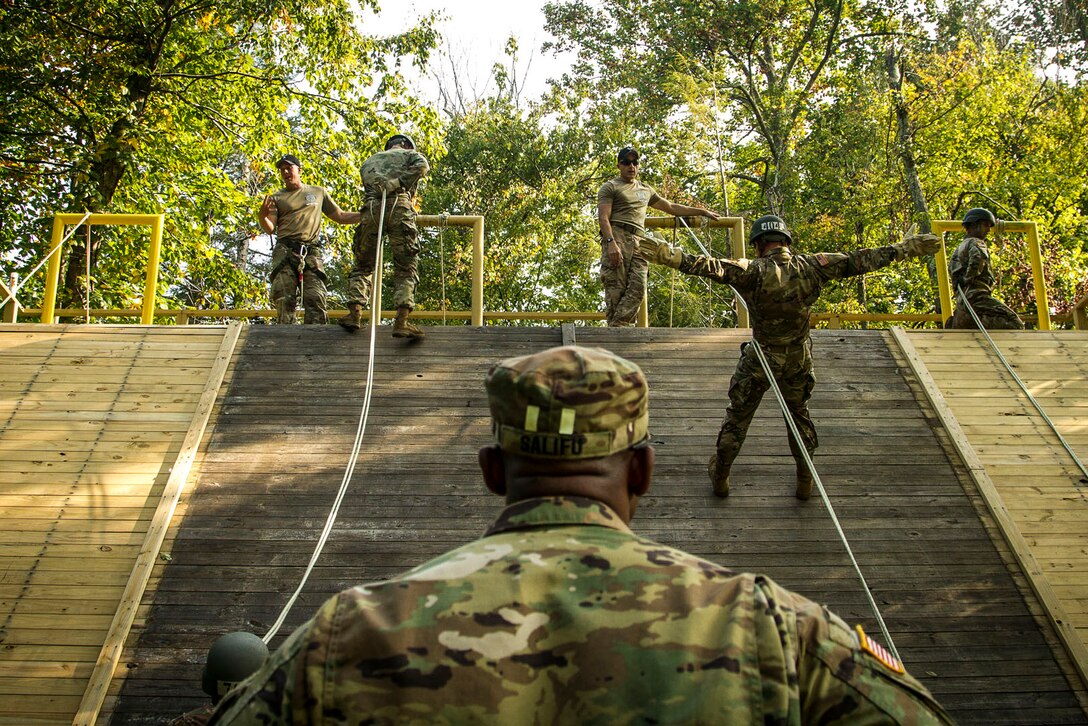 Soldiers walk rappel down a wall as instructors observe at the top and on the ground.