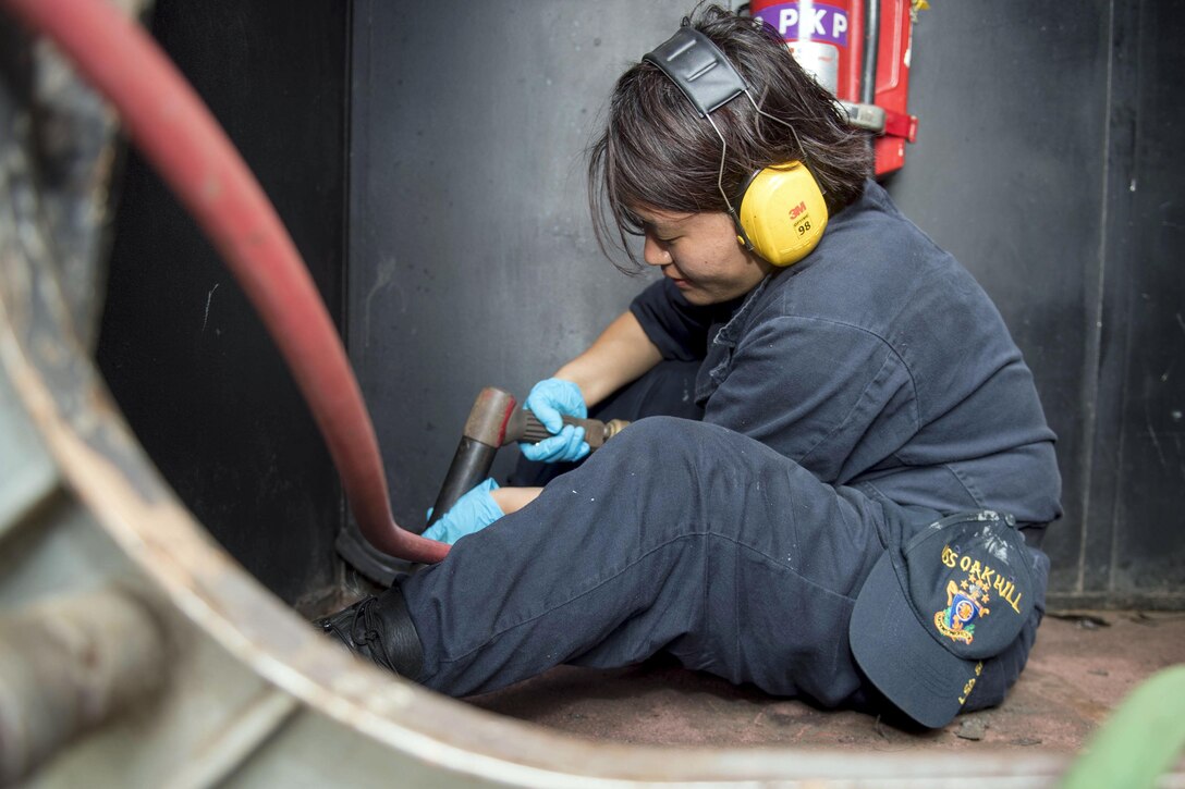 A sailor wearing headphones sits on a ship floor and operates a needle gun.
