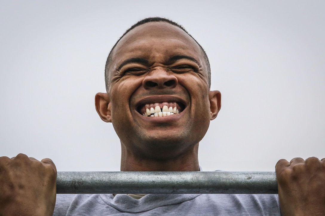 An airman clenches his teeth while hanging from a bar and holding his head over it.