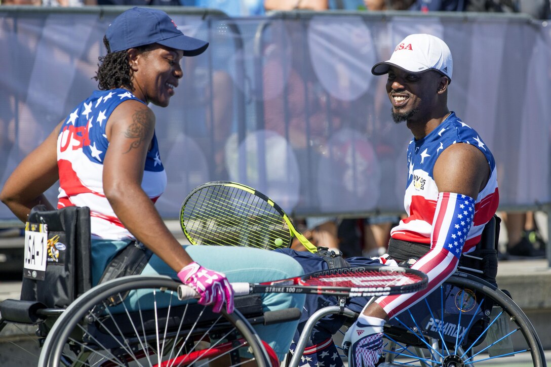 Two athletes in wheelchairs holding tennis rackets look at each other.