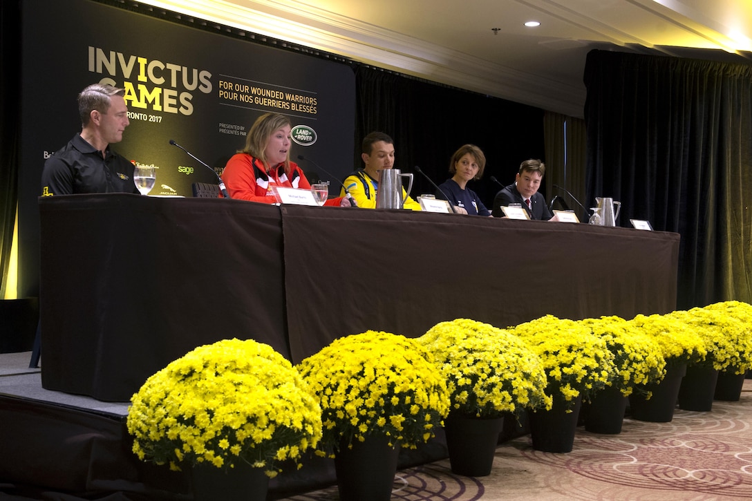 A row of five people sit at a table with a sign that says Invictus Games behind them.