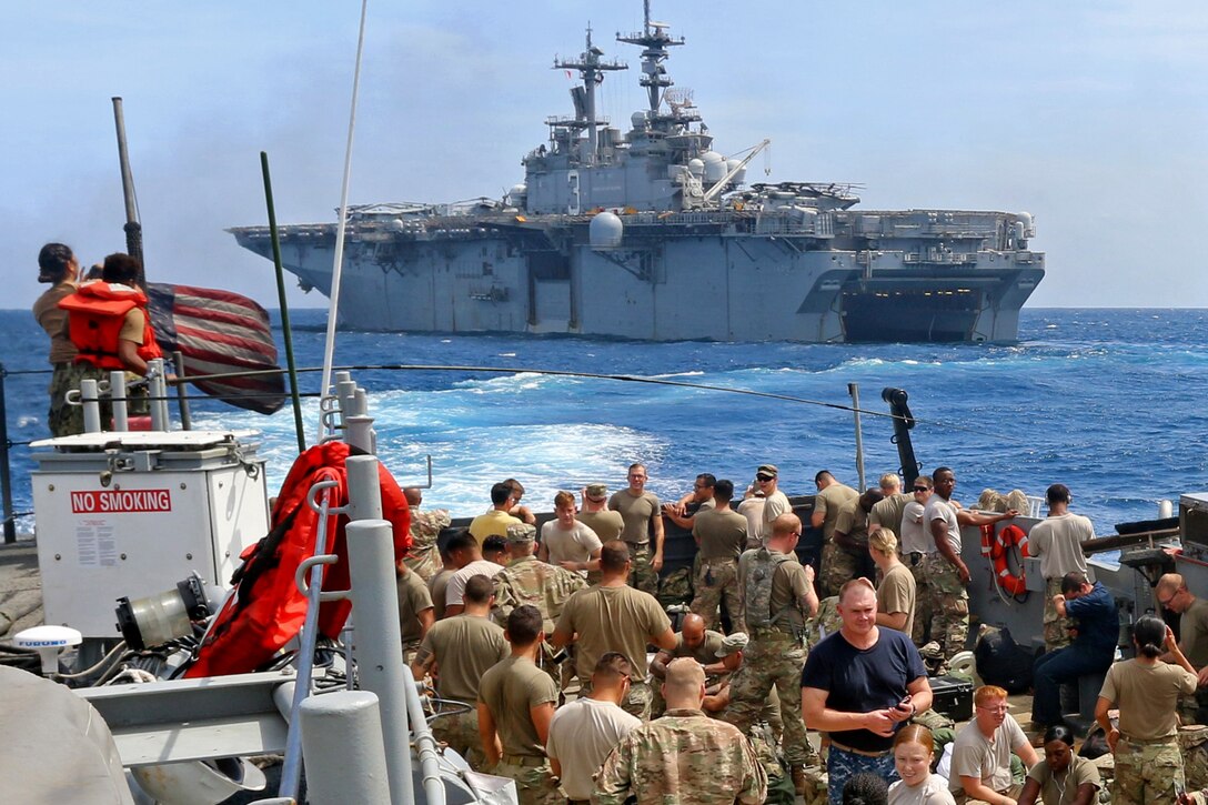 Service members stand on the deck of one ship looking at another ship.