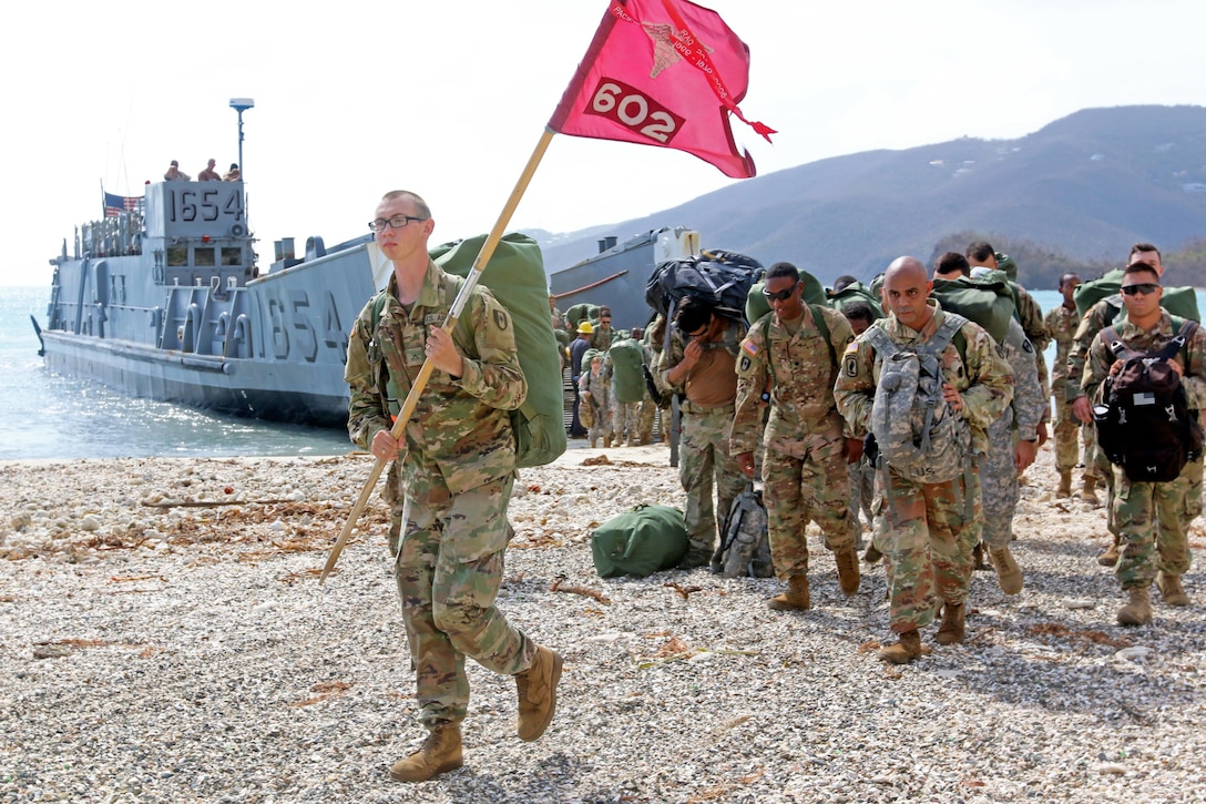 A soldier carrying a flag leads other soldiers off a boat.