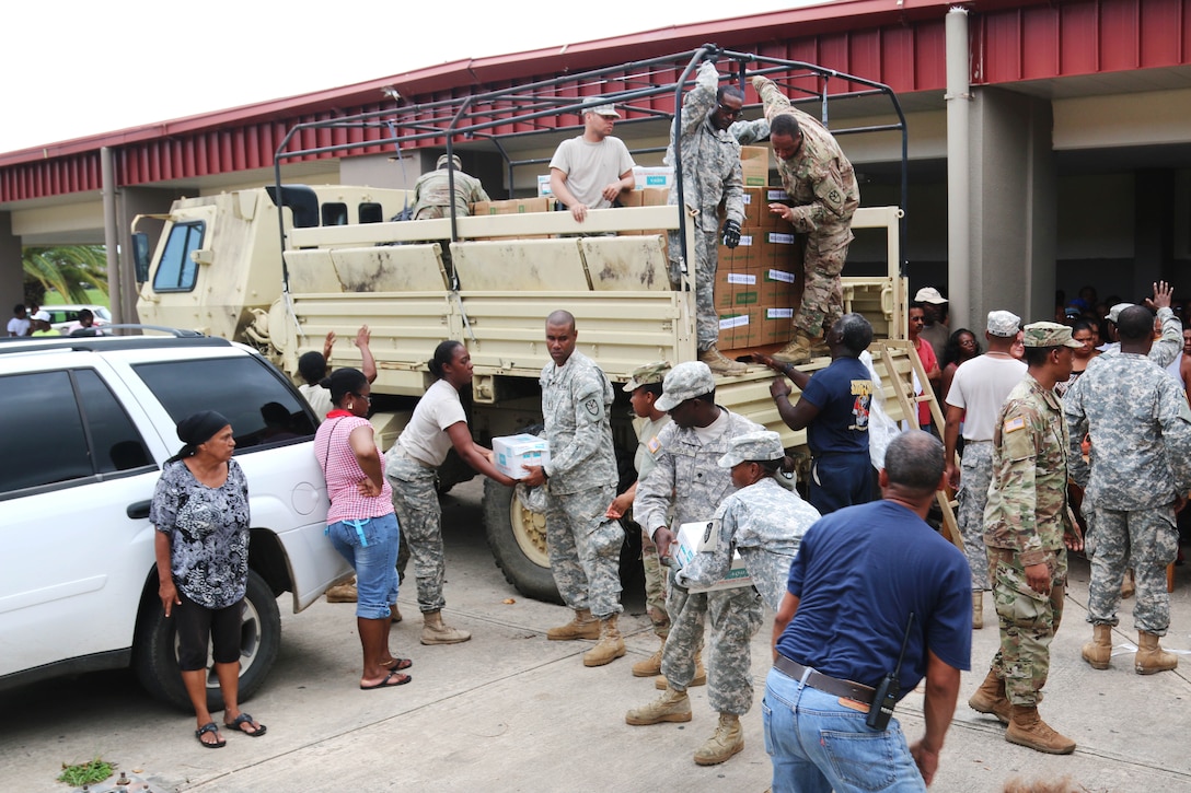 Soldiers distribute food and water off the back of a military vehicle.