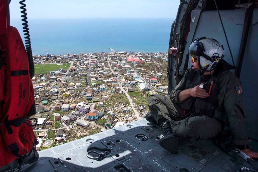 A sailor looks out the open door of a helicopter in flight with buildings and water below.