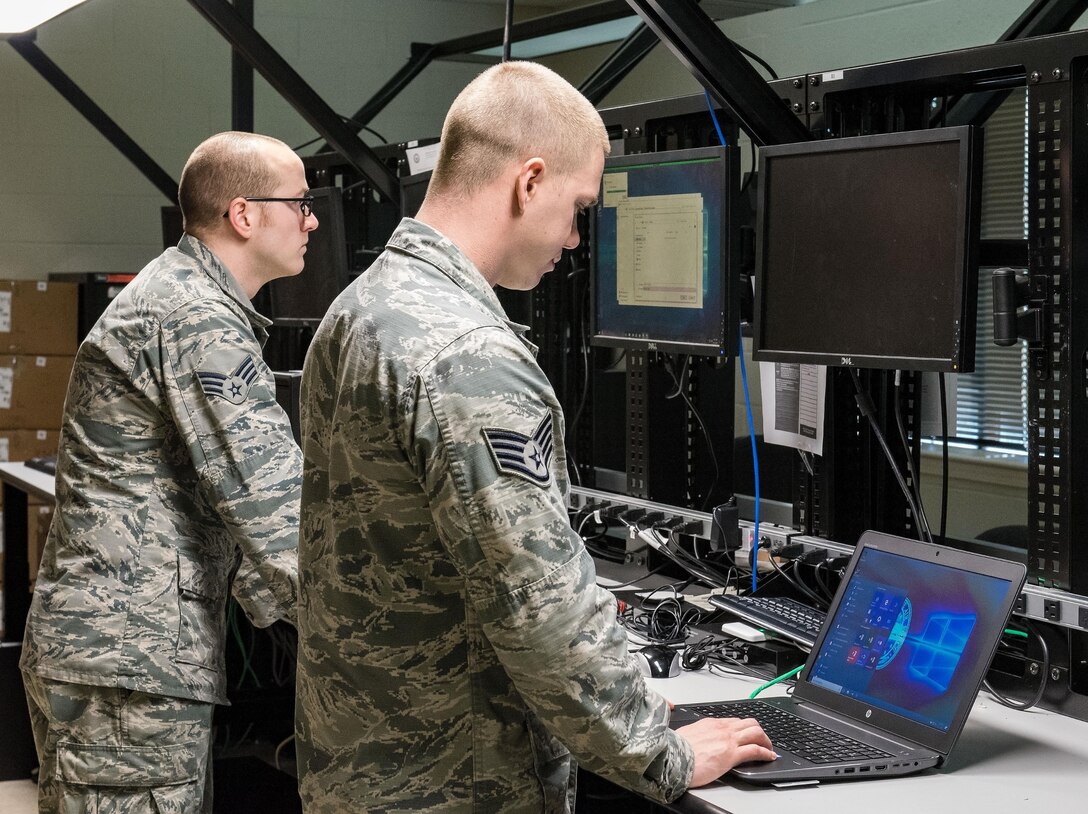 Senior Airman Tyler Scoble, 436th Communications client service technician, left, and Staff Sgt. Eric Rzepa, 436th CS Comm Focal Point NCO in charge, center, work on upgrading a desktop and laptop computer on the consolidated work bench Sept. 21, 2017, on Dover Air Force Base, Del. CST and CFP personnel, and unit CSTs with administrative privileges have the capability to rollout up to 250 assets per week with Windows 10 Secure Host Baseline version 5.3. (U.S. Air Force photo by Roland Balik)