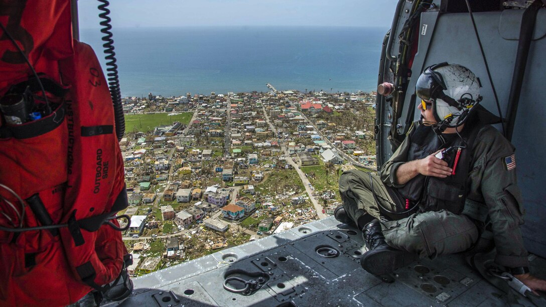 A sailor looks out from an open helicopter door over a coastal area dotted with damaged and destroyed buildings.