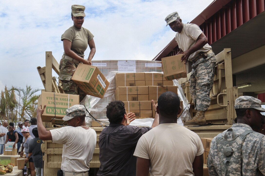 Soldiers standing in the cab of a military truck pass cardboard boxes to troops and civilians on the ground.