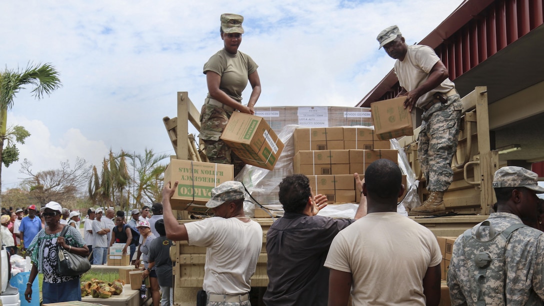 Soldiers standing in the cab of a military truck pass cardboard boxes to troops and civilians on the ground.