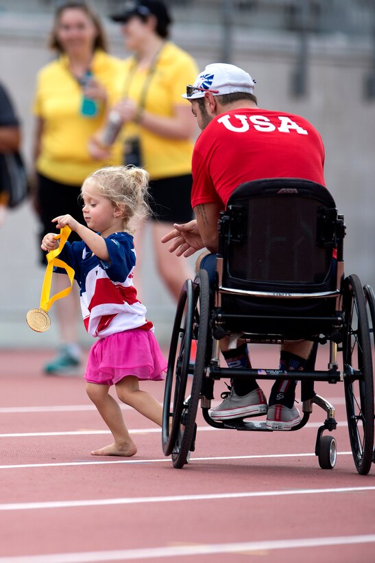 A 3-year-old girl runs while holding up her father’s gold medal .