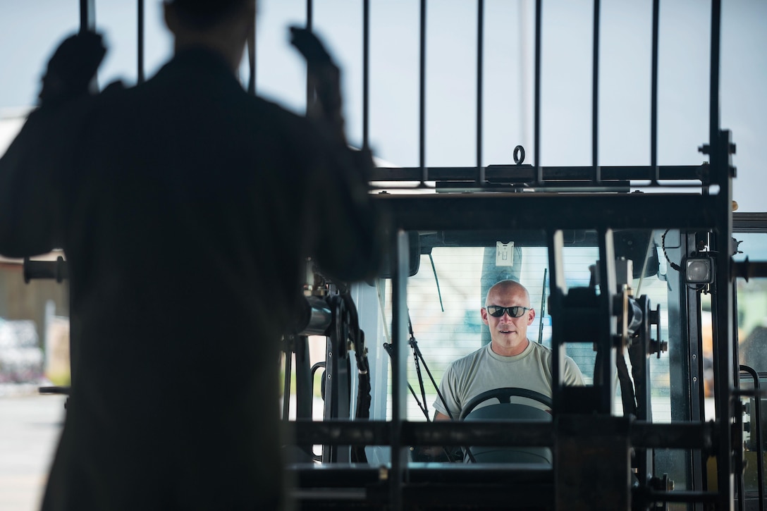 An airman gives hand signals to another airman in a forklift.