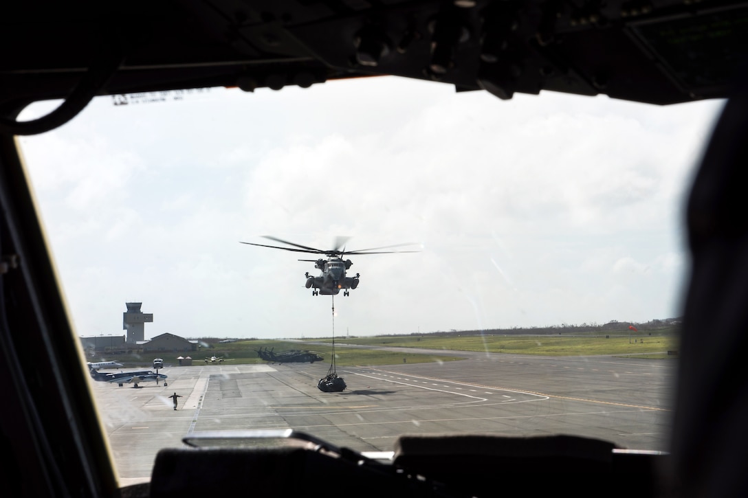 A helicopter in the air lifts cargo attached a the end of a line.