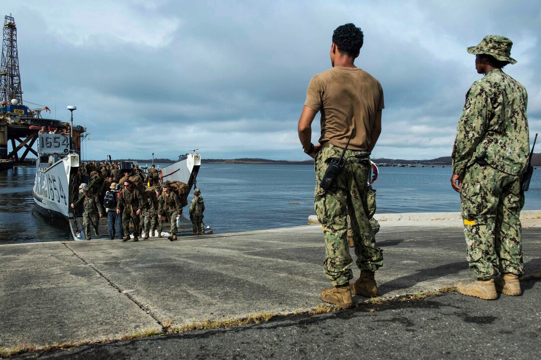 Marines walk off a landing craft as two service members observe from a distance.