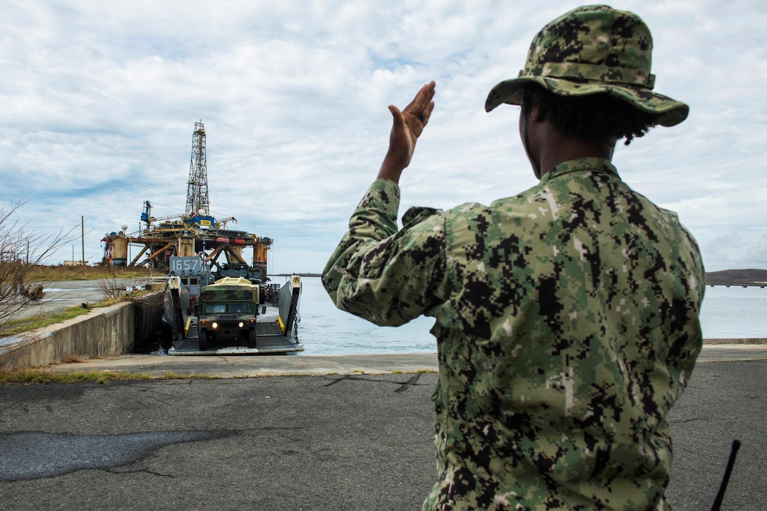A sailor signals to a Humvee driving off a landing craft.