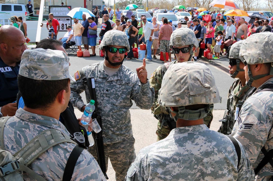 A soldier talks to other soldiers and residents stand in the background.