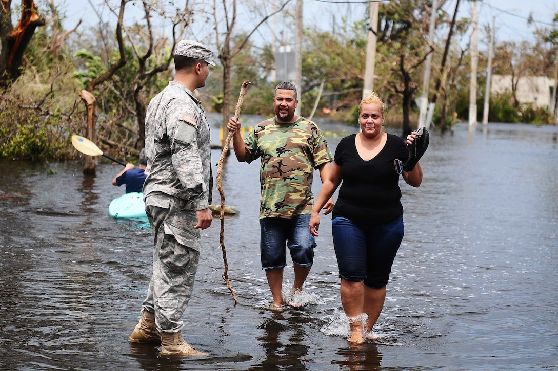 A soldiers helps a man and a woman through a flooded area.