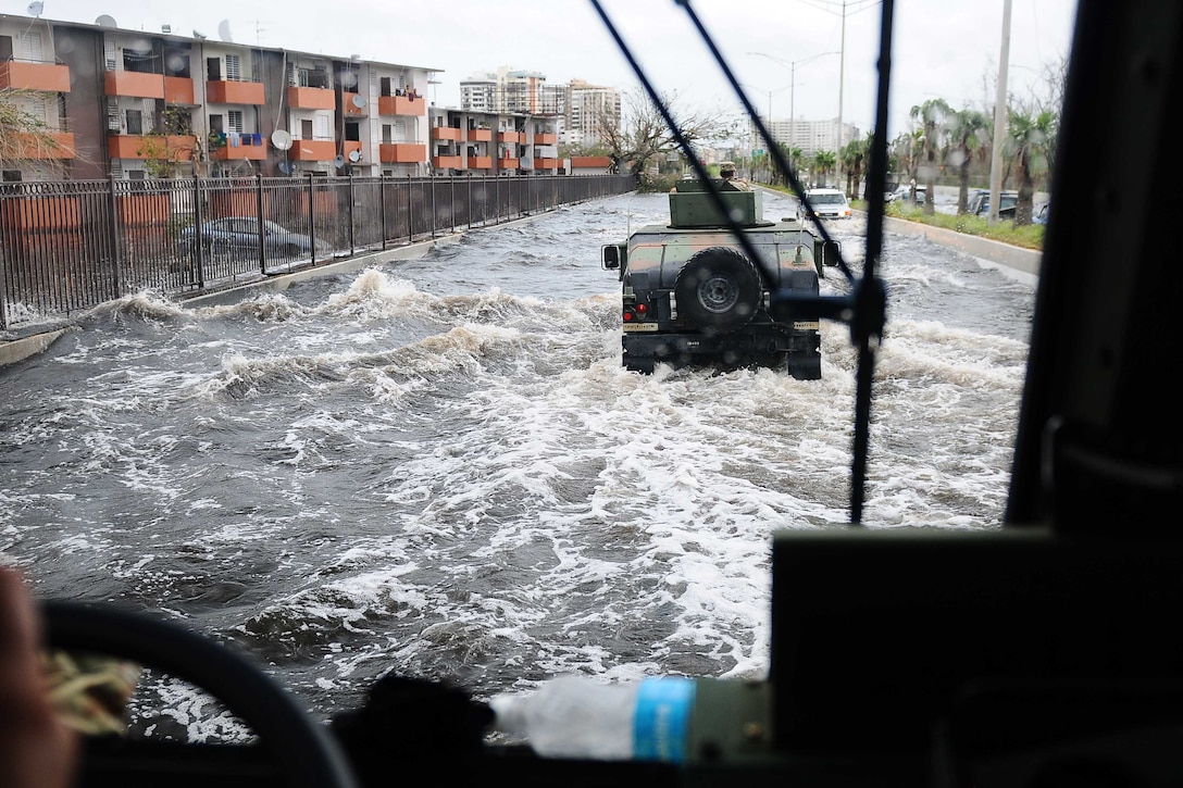 Military vehicles drive through a flooded highway.