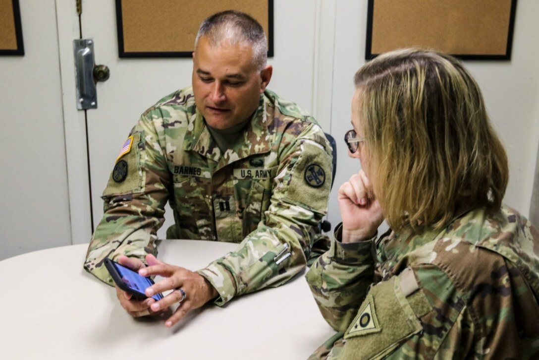 Two service  members sit at a table looking at a cell phone.