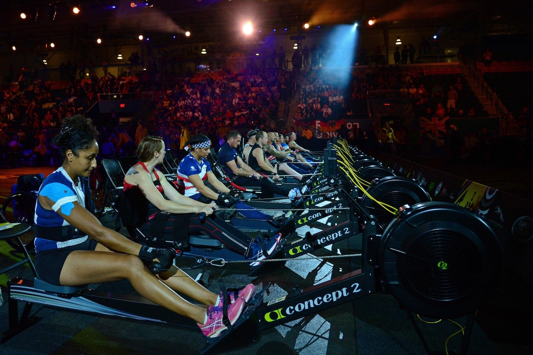 A line of athletes sit at indoor rowing machines with people watching from the stands.