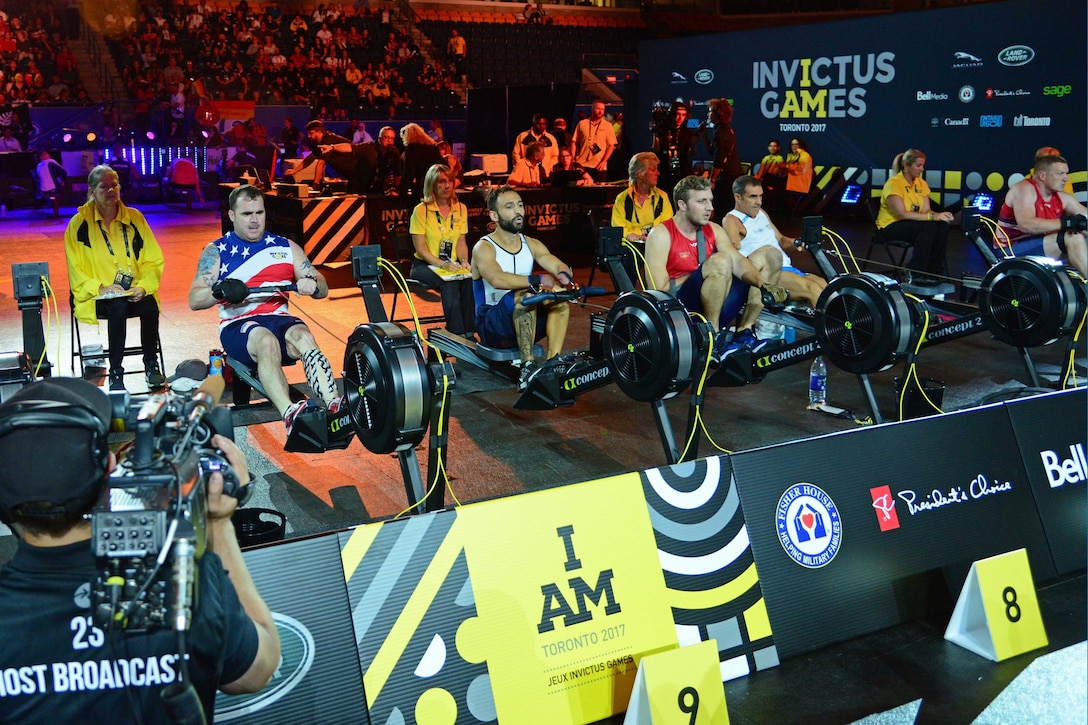 A line of people use rowing machines in a stadium.