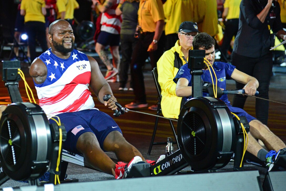 Two people use rowing machines with people standing behind them.