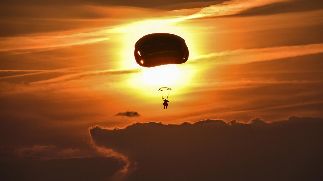 A soldier, shown in silhouette, parachutes toward the ground against an orange sky.
