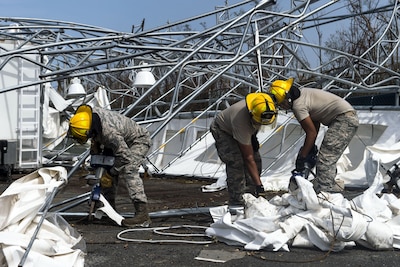 Troops in hard hats clean up debris on the ground near mangled metal structures.