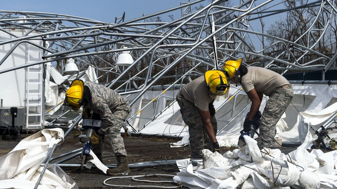 Troops in hard hats clean up debris on the ground near mangled metal structures.