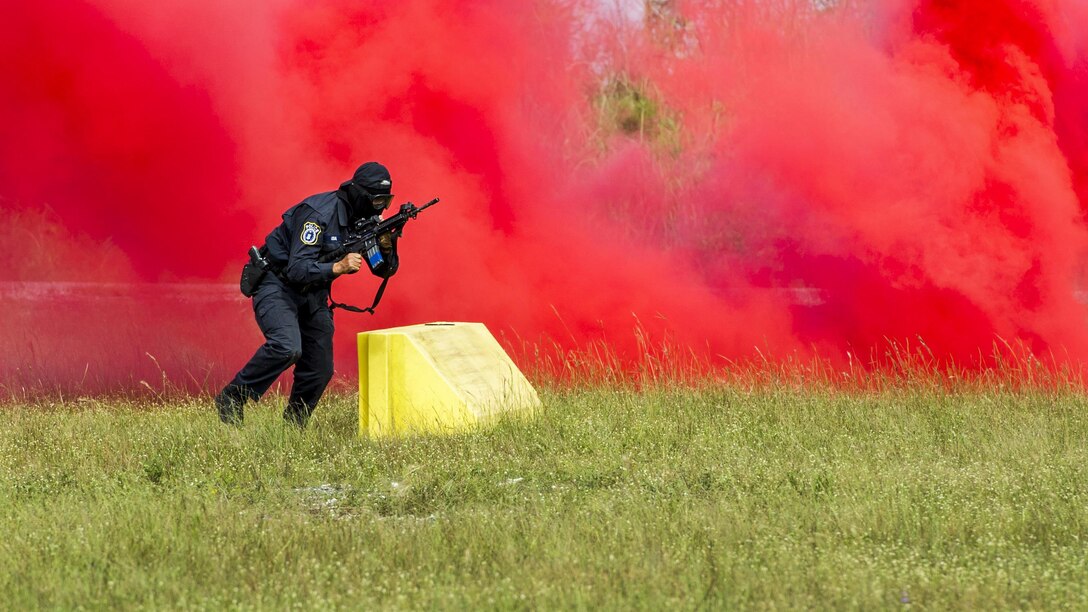 An airman moves with a weapon in front of a red cloud of smoke.
