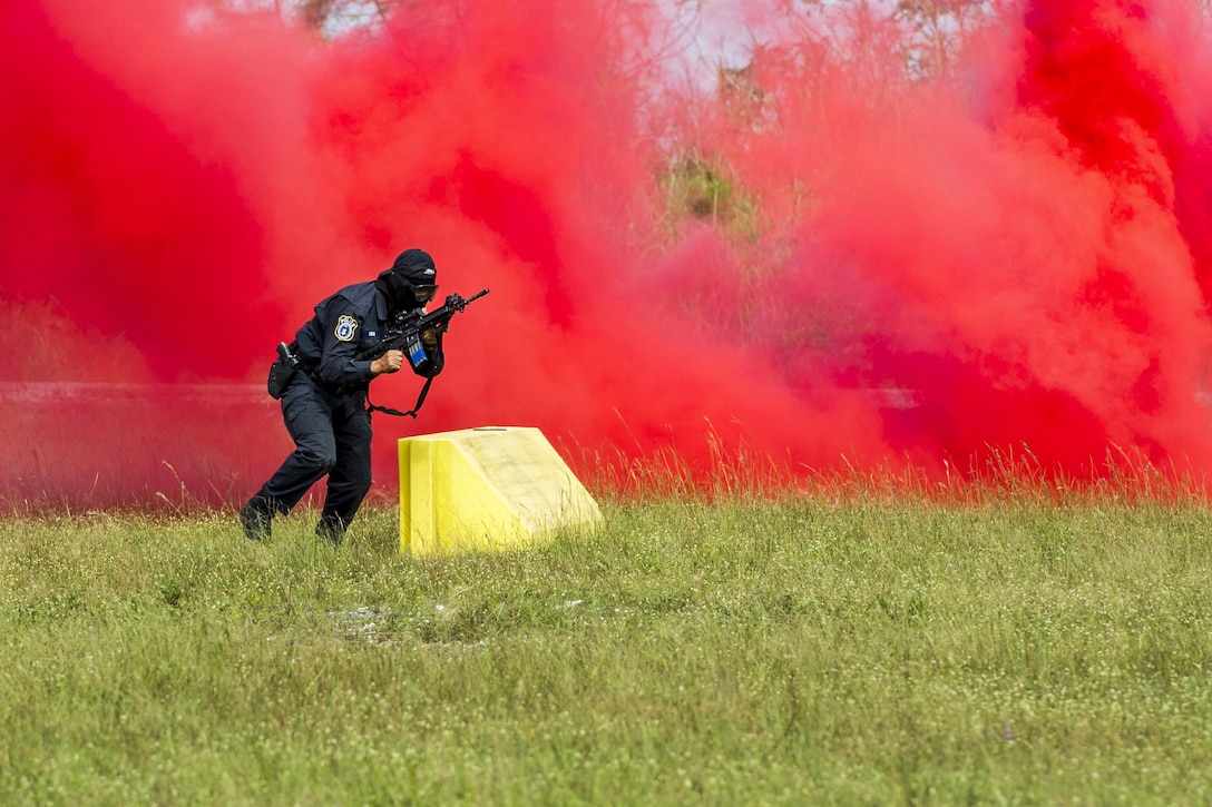 An airman moves with a weapon in front of a red cloud of smoke.